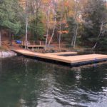 A floating wooden dock on a calm lake surrounded by trees with autumn foliage, and a small platform with a bench on the shore.