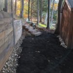 A shaded path with fresh mulch nestled between a wooden shed and a wooden retaining wall, scattered with stones and a few boxes.