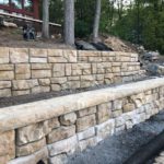 Newly constructed retaining walls made of natural stone in a landscaped area with a house and trees in the background.