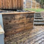 Wooden outdoor shower enclosure with a bench, situated on a wooden deck surrounded by autumn leaves.