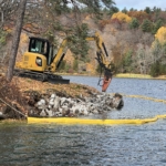 An excavator with a grapple attachment working on a rocky shoreline, surrounded by a yellow containment boom on a lake during autumn.