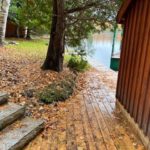 Wet wooden path with fallen leaves alongside a cabin, leading to a lake with visible green boats, surrounded by trees.