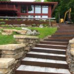 Stone steps leading up to a house with large windows and a red pergola, with a yellow excavator parked beside the house.