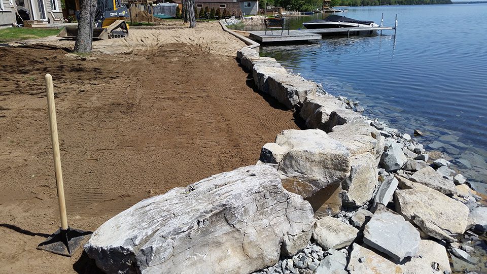 A shoreline area under construction with large rocks forming a barrier, freshly laid sand, a shovel upright in the ground, and a dock extending into the water.