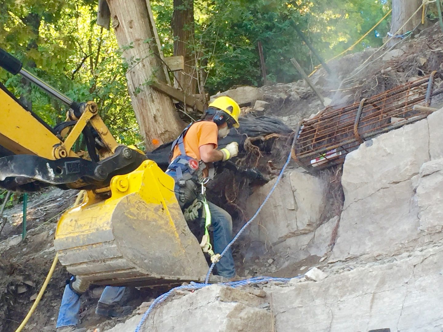 Construction worker in safety gear using heavy machinery for stabilization work on a steep, rocky shoreline.