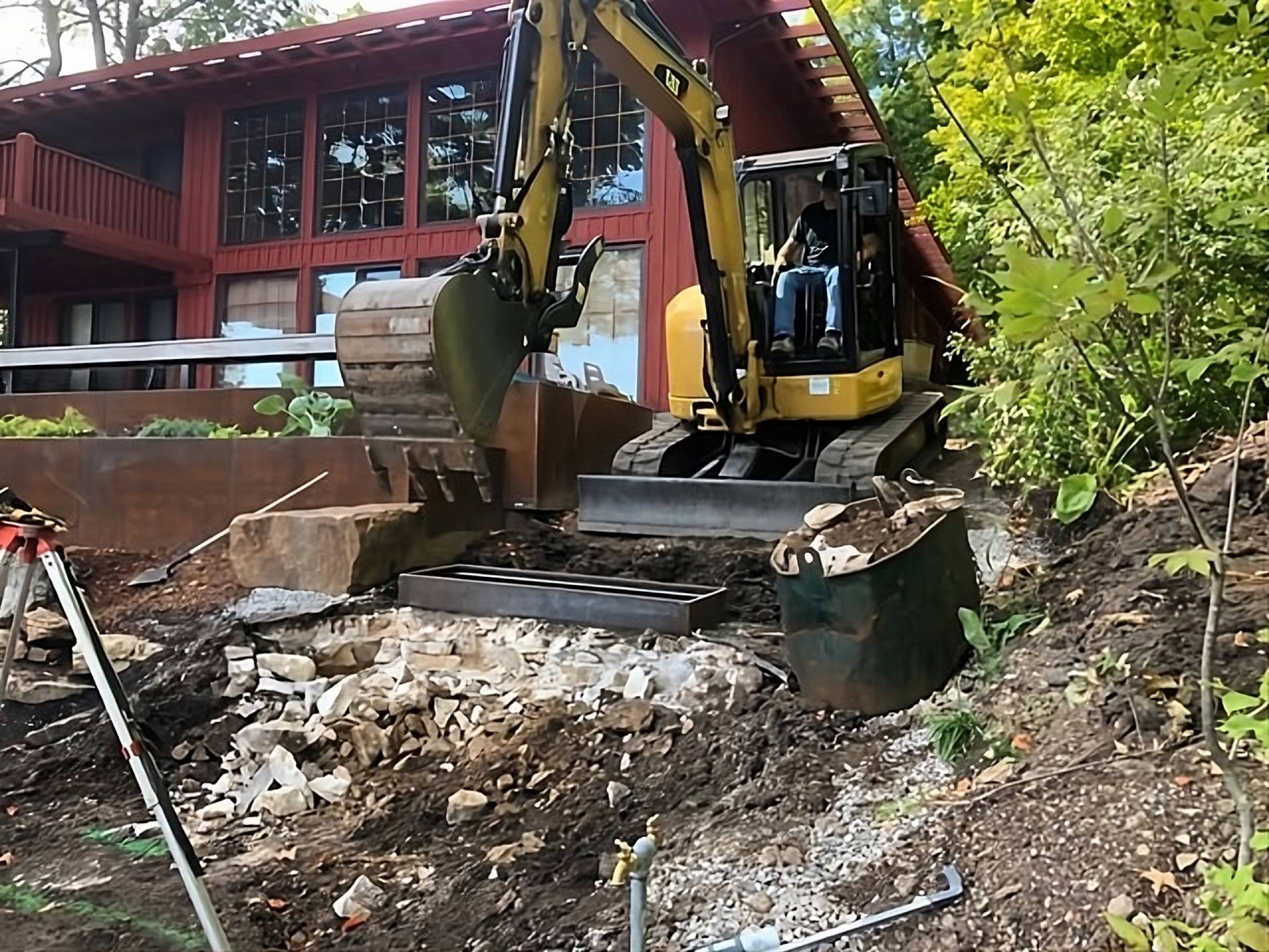 An excavator operating in a residential area, digging near a red house surrounded by trees and debris.