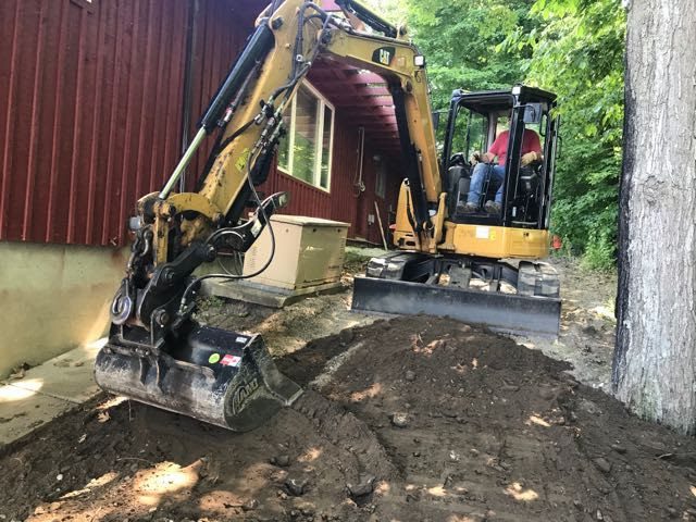 A person operates a yellow excavator near a red metal building, digging and moving dirt. trees in the background suggest a wooded area.