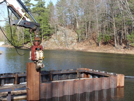 A construction crane operating near a metal cofferdam along the river shoreline, with trees in the background.