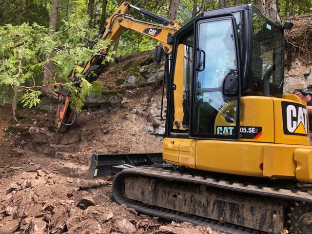 A yellow caterpillar excavator with a circular saw attachment clearing shrubs near the docks.