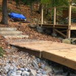 Wooden walkway and steps in a forested area with scattered leaves, leading to a deck and covered storage area.