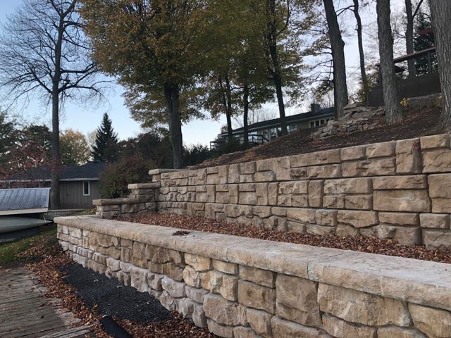 Dry-stacked stone retaining walls with stair steps in a park-like setting, surrounded by trees, boathouses, and a distant bridge.