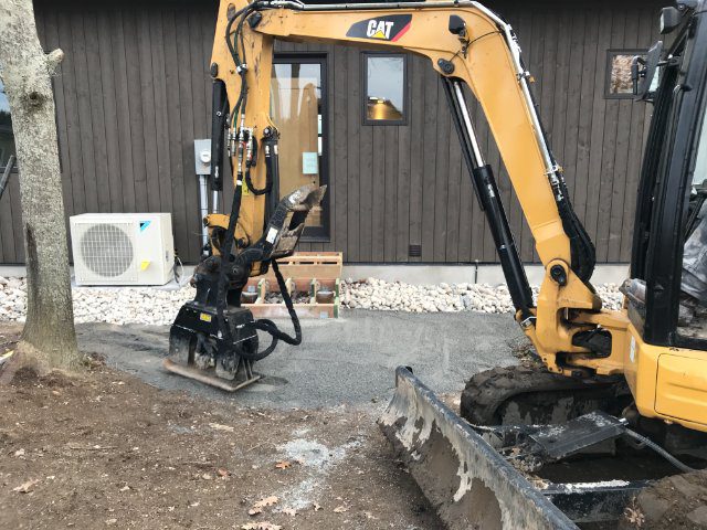 A yellow excavator parked in front of a brown building, with a white heat pump unit visible to the side.