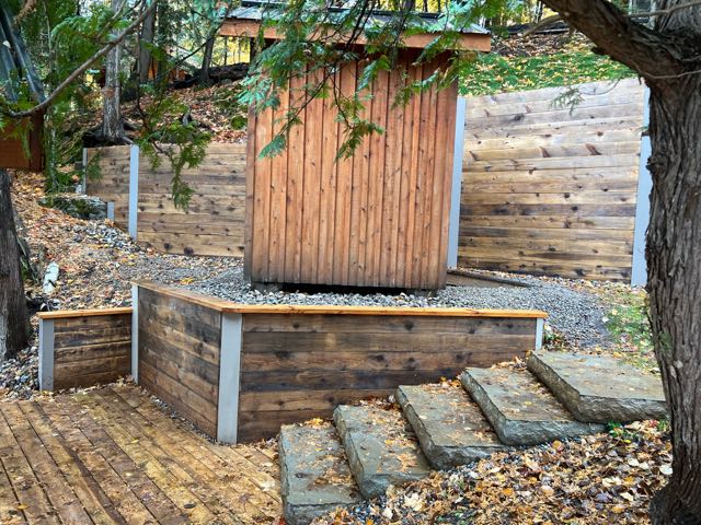 Wooden backyard compost bin surrounded by trees, with a stone pathway leading to it, set against a fence near boathouses.