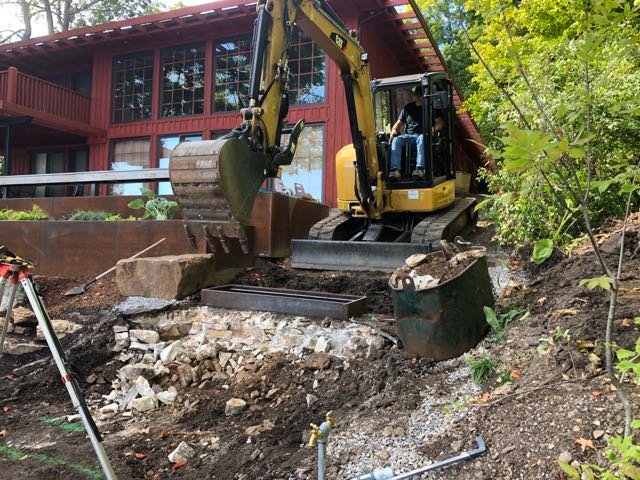 A yellow excavator working on a rocky hillside near a red house, with large metal retaining walls partially installed.