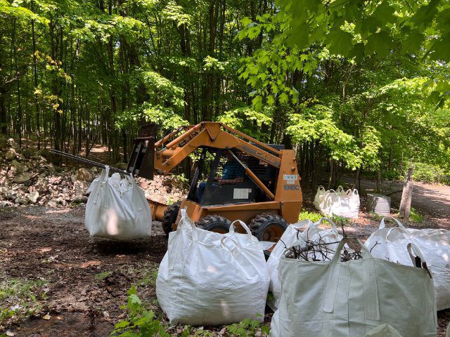 A skid steer loader moving large white bulk bags at the docks in a forested area.