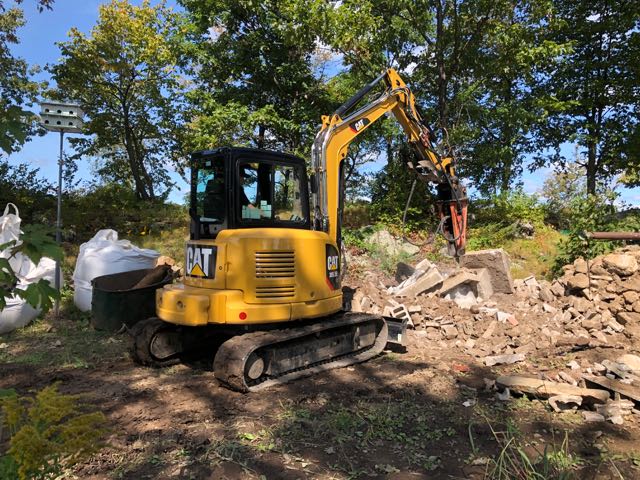 A yellow caterpillar excavator parked on a rubble-strewn site with trees in the background.