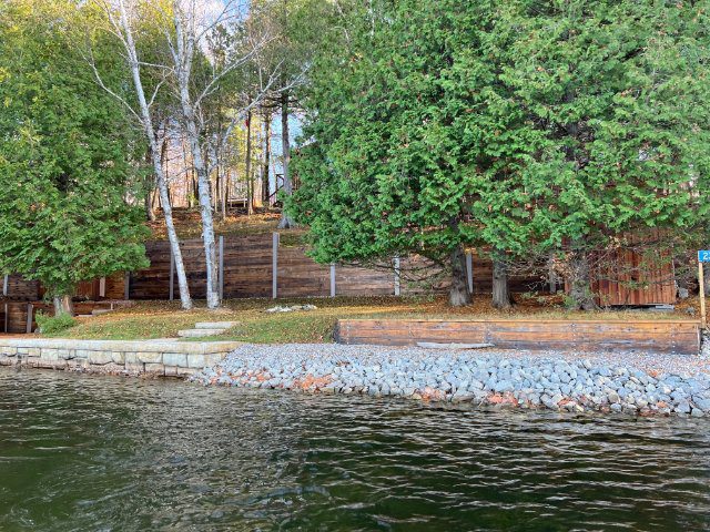 A tranquil lakeside scene with a rocky shore in the foreground and a wooden boathouse partially obscured by birch and evergreen trees.