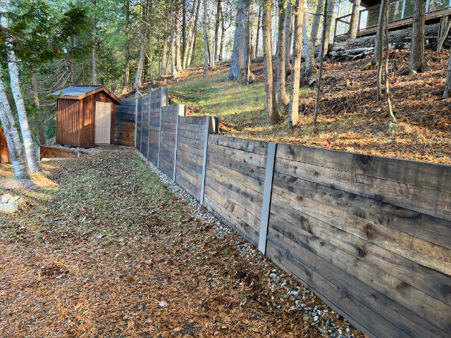 Wooden fence leading to a small cabin in a forested area with sunlight filtering through the trees, near secluded boathouses.