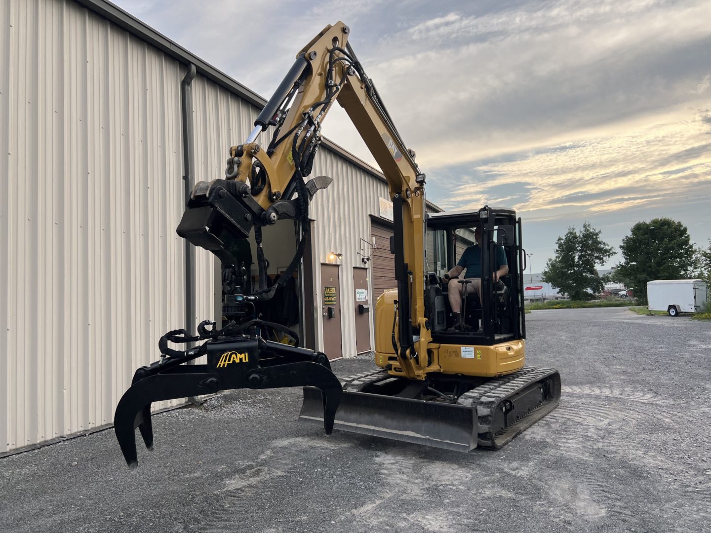 A yellow excavator with a log grapple attachment parked beside a warehouse, with a driver in the cabin.