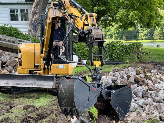 A man operating a yellow excavator with an attached grapple, surrounded by rocks and greenery near a pond.