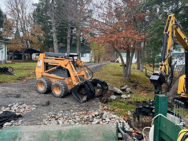 A skid steer loader and an excavator working on a residential gravel driveway with scattered piles of stones and debris, surrounded by trees and a white house.