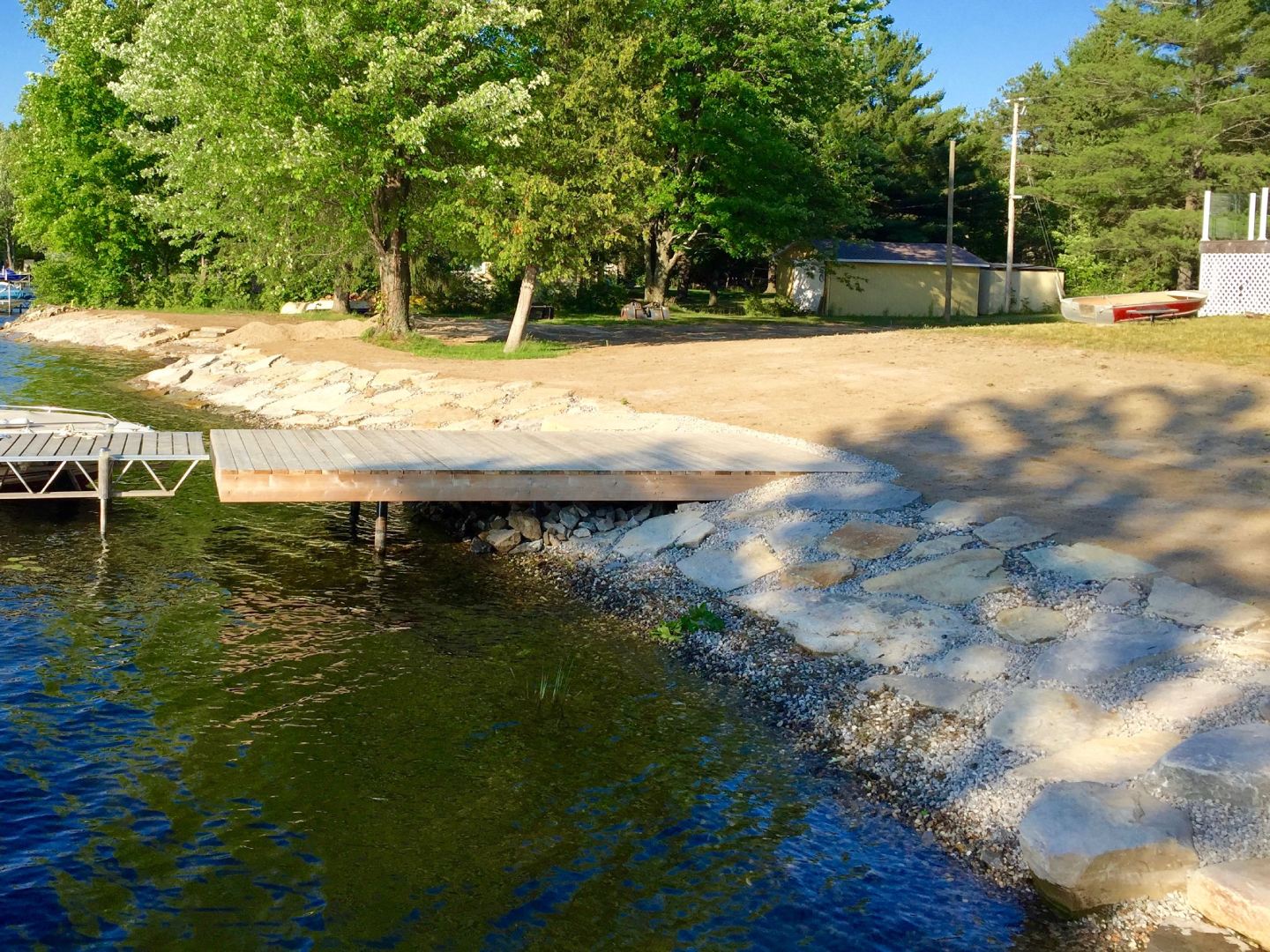 Wooden dock extending into a calm lake with surrounding trees and a rocky shoreline under a clear sky.